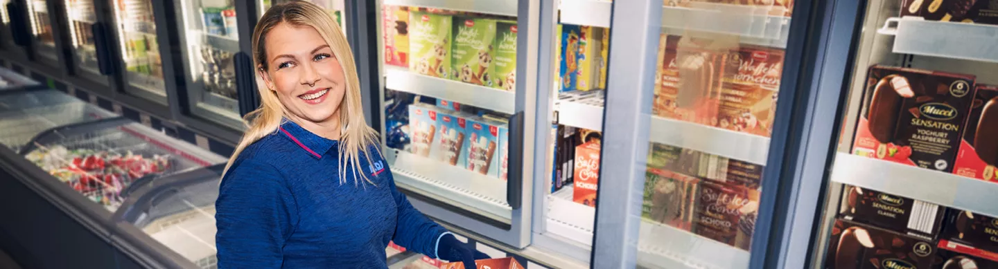 Eine lächelnde Mitarbeiterin in ALDII-Arbeitskleidung steht im Markt vor Kühlregalen und befüllt diese mit Ware. 