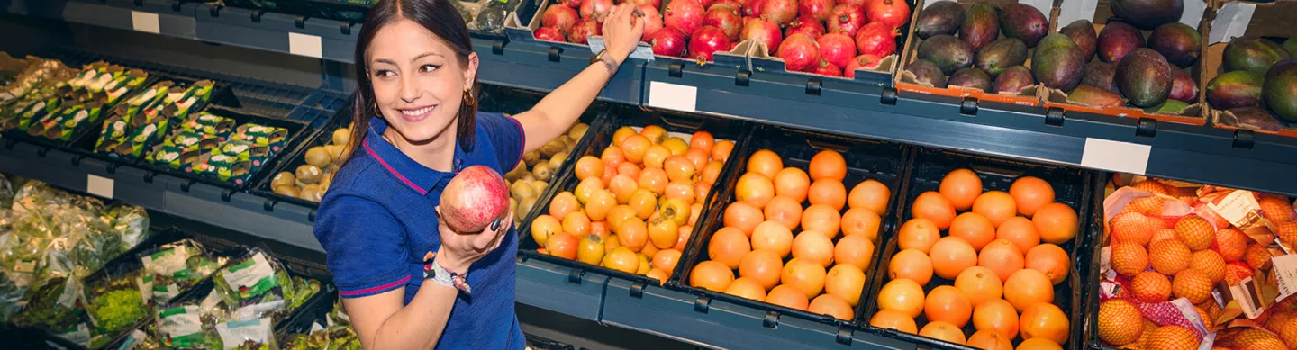 Verkäuferin steht in einem Supermarkt vor der Obst- und Gemüseabteilung und lächelt in die Kamera. In der Hand hält sie einen Granatapfel.