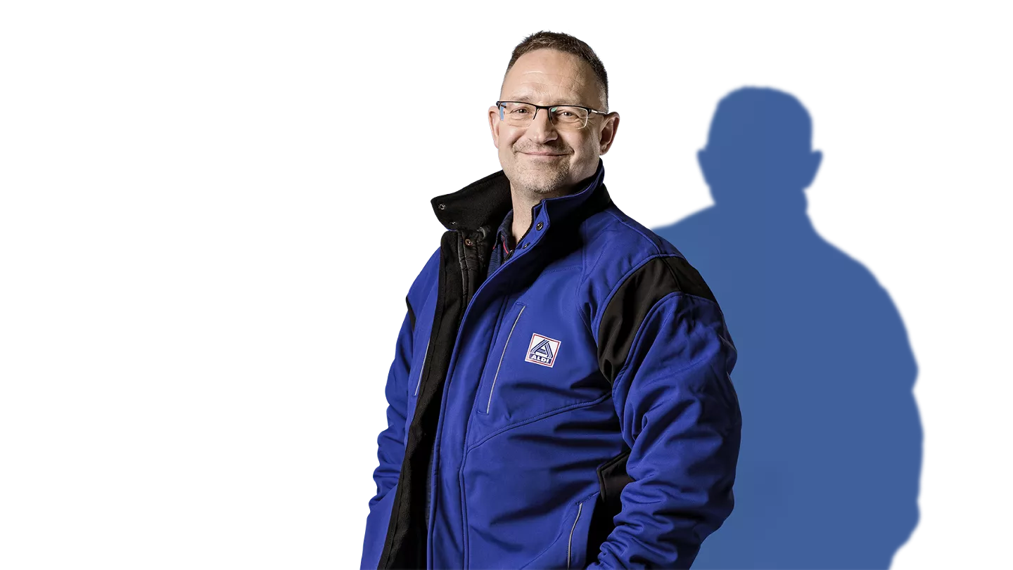 Ein Mann mit ALDI Poloshirt und blau-gelber Warnweste. Er lächelt leicht und hält die Weste auf Brusthöhe fest.