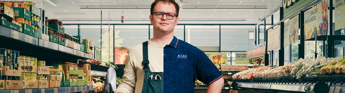Mitarbeiter von ALDI Nord steht selbstbewusst in einer Filiale zwischen Regalen mit Lebensmitteln. Er trägt Arbeitskleidung mit ALDI Nord Logo und hält eine kleine Schaufel in der Hand. Im Hintergrund sind Obst- und Gemüseregale sowie Verkaufsflächen zu sehen