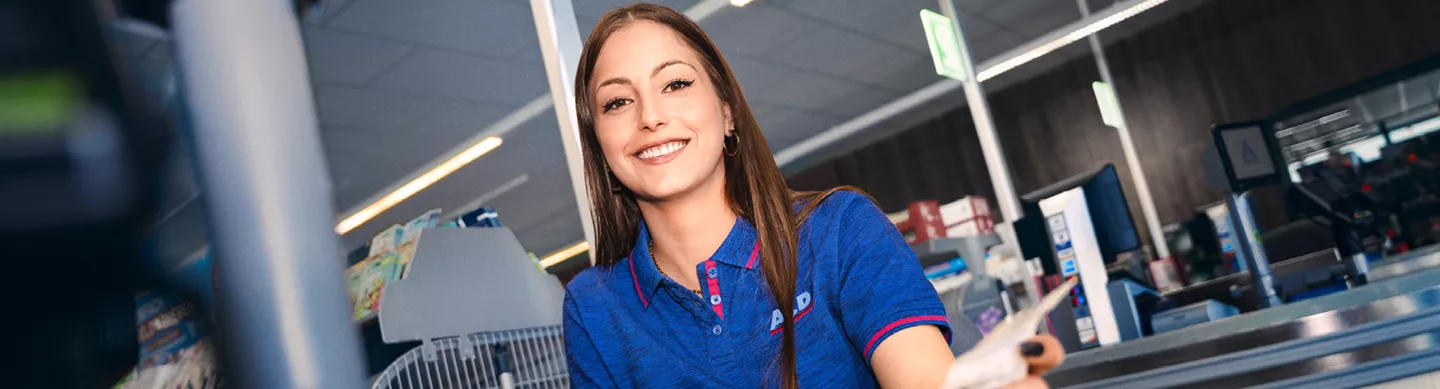 Mitarbeiter von ALDI Nord steht selbstbewusst in einer Filiale zwischen Regalen mit Lebensmitteln. Er trägt Arbeitskleidung mit ALDI Nord Logo und hält eine kleine Schaufel in der Hand. Im Hintergrund sind Obst- und Gemüseregale sowie Verkaufsflächen zu sehen