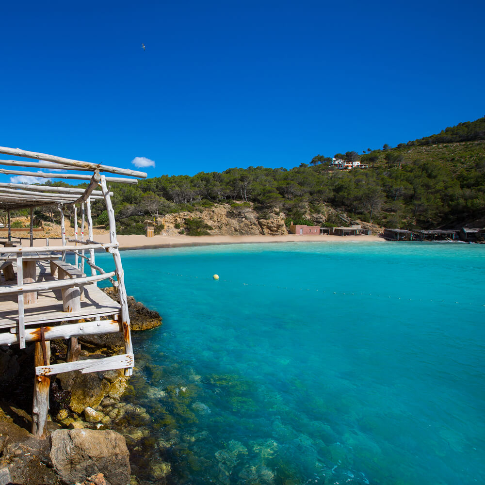 Benirrás Beach: Topaz rocky sea,  a white wooden structure next to it and the beach in the distance