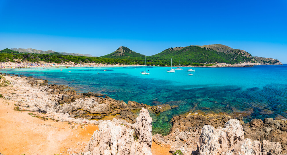 Cala Agulla: Golden cliffs and blue ocean dotted with boats with cliffs in the background
