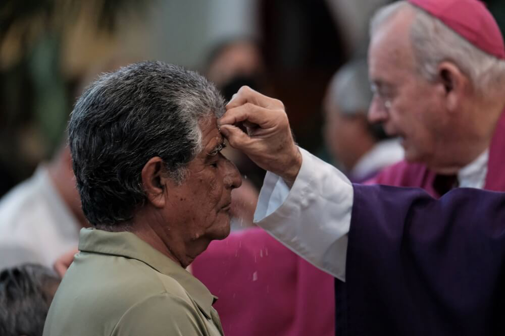 A close-up photo of a priest marking the sign of the cross on a man’s forehead 