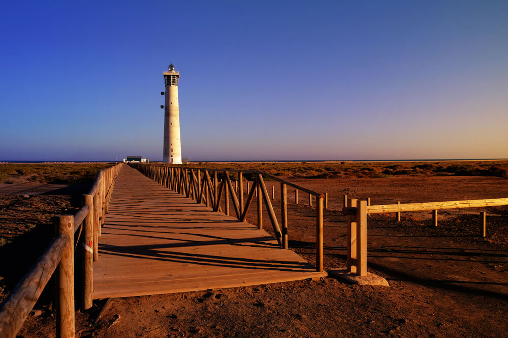 Fuerteventura-Urlaub: Steg zu einem Strand mit Leuchtturm und Vegetation.