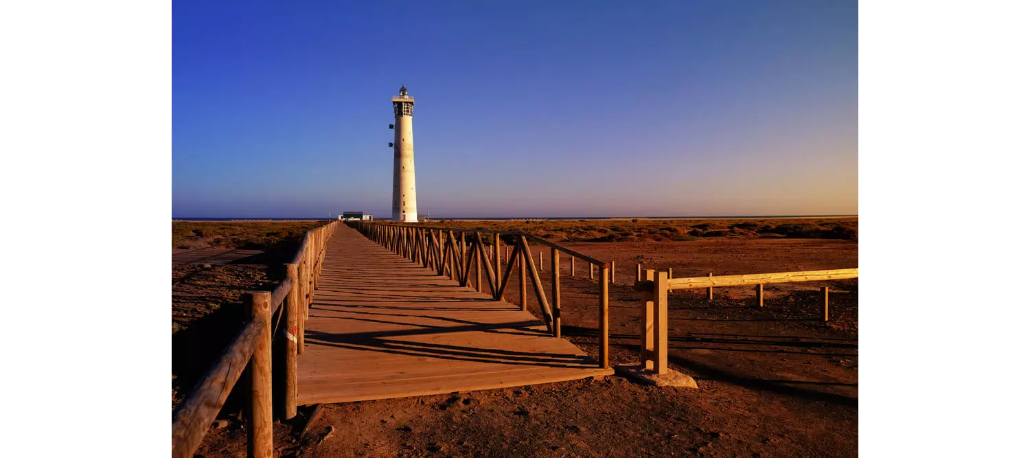 Fuerteventura-Urlaub: Steg zu einem Strand mit Leuchtturm und Vegetation.