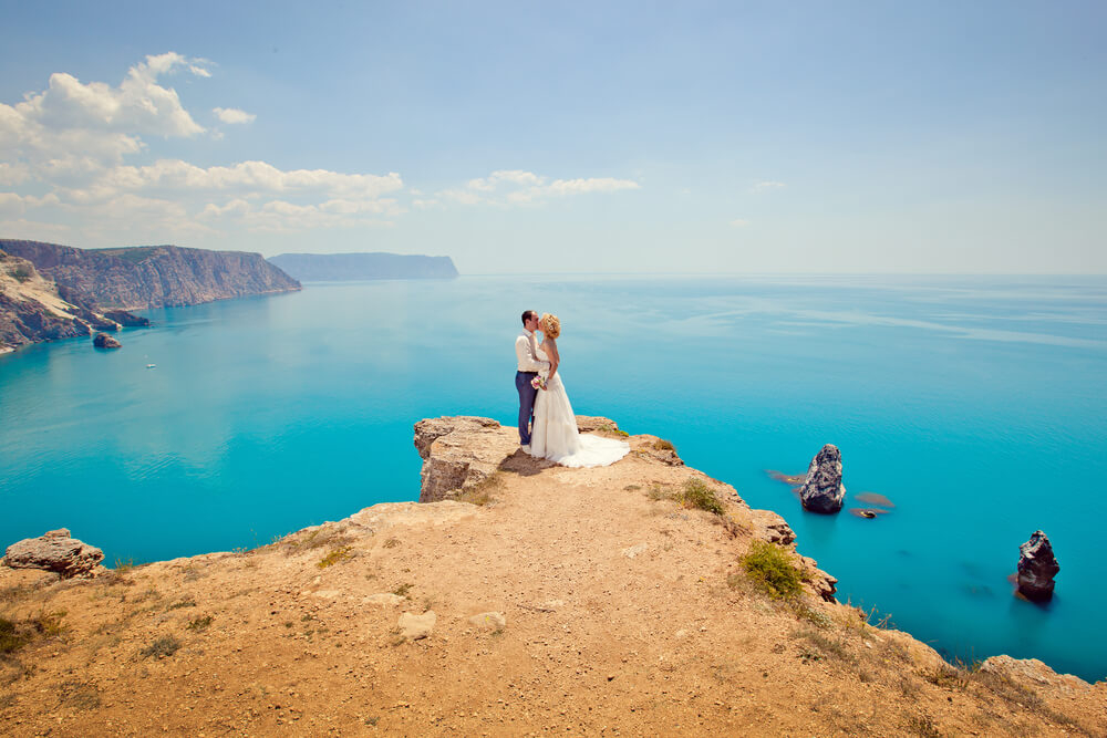  Hochzeit im Freien: Brautpaar an einer Klippe mit blauem Meer und weiteren Klippen im Hintergrund