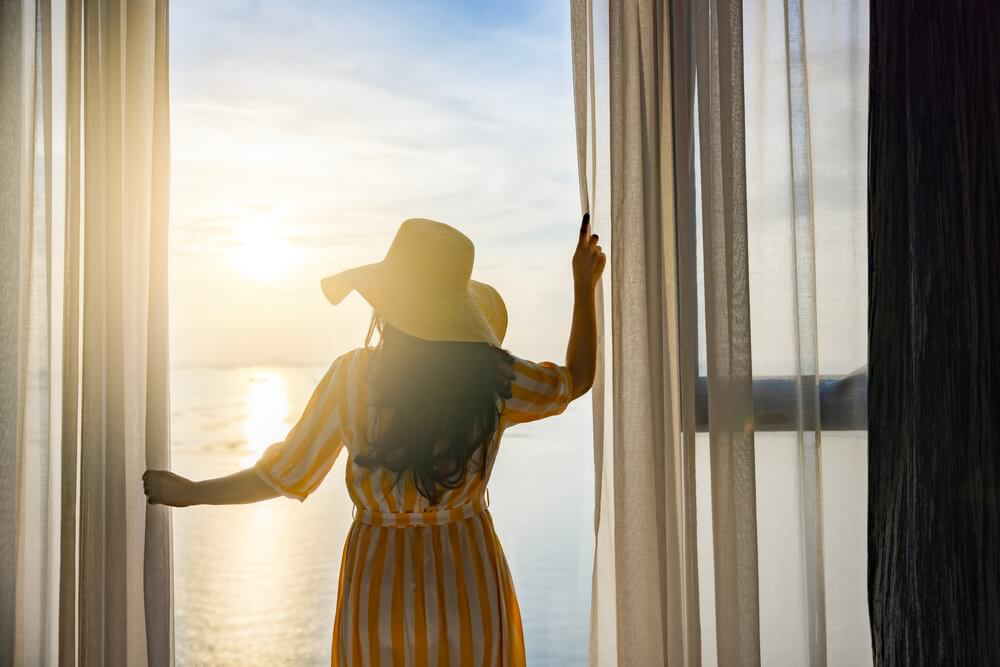 Frau öffnet Hotelbalkon zum Sonnenaufgang: für den Urlaub ist die Zeitumstellung sinnvoll.