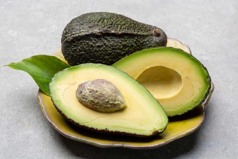 A large Mexican avocado, cut in half, sits on a yellow plate.