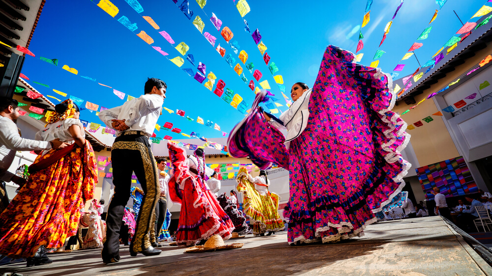Folk dancers wearing colorful Mexican clothing dance among colorful banners hanging overhead.