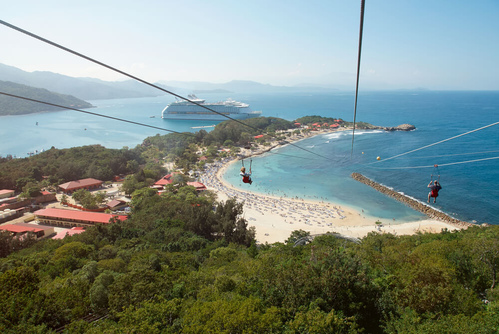 Two people zipline over views of a beach and the Caribbean Sea.