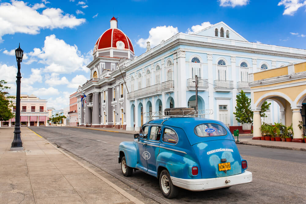 Cienfuegos: A vintage blue Ford car parked in front of pastel coloured buildings