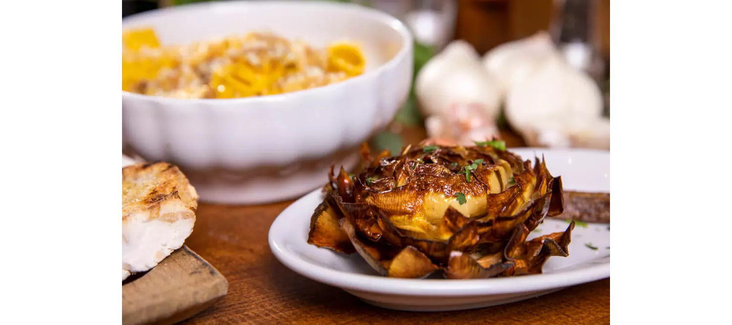 Carciofi alla Giudia: A rose shaped artichoke roasted on a white plate on a wooden table
