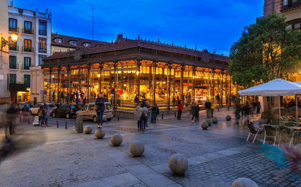 Mercado San Miguel Madrid: A Madrid street at night with a glass-walled market building