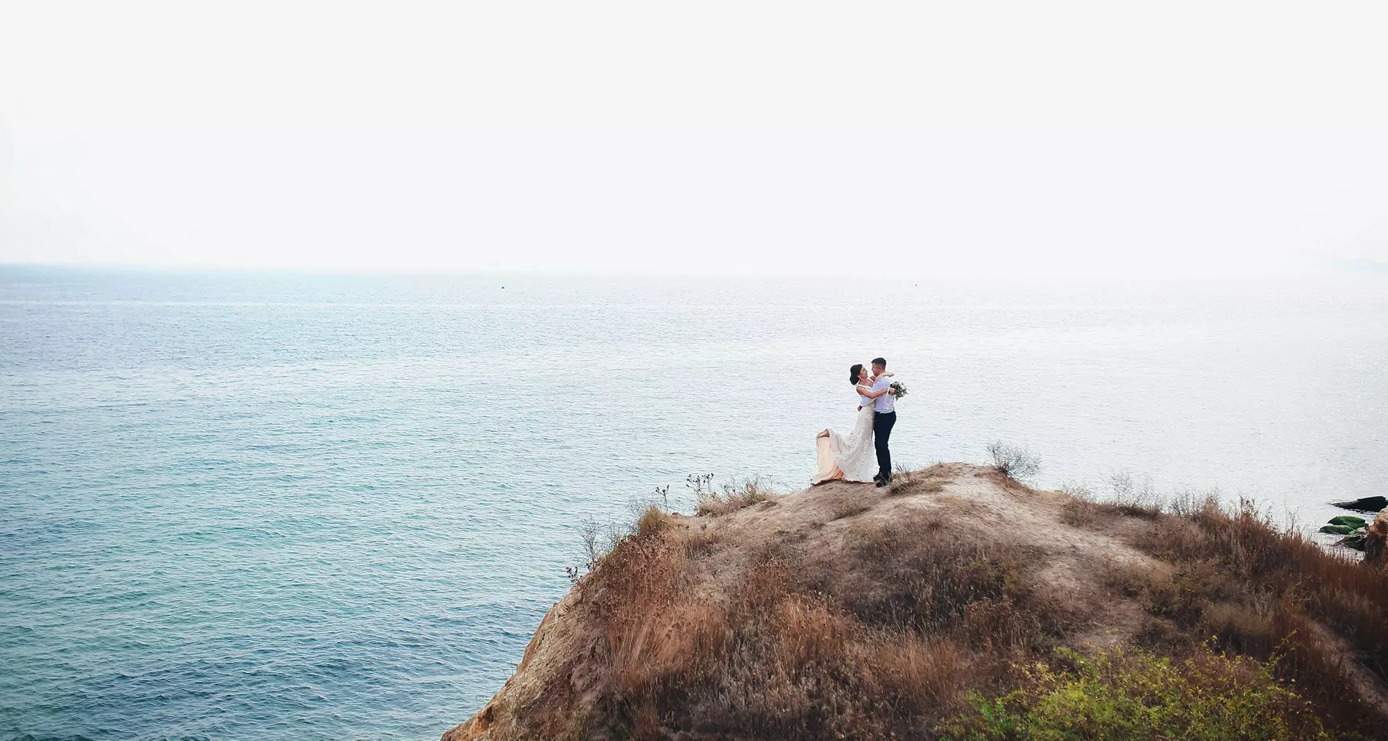 Wedding Destinations: Married couple on a cliff hugging with the sea in the background