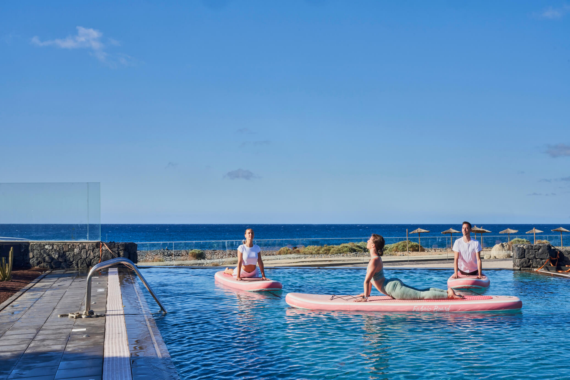 Three people practice paddleboard yoga the pool on their yoga retreat.