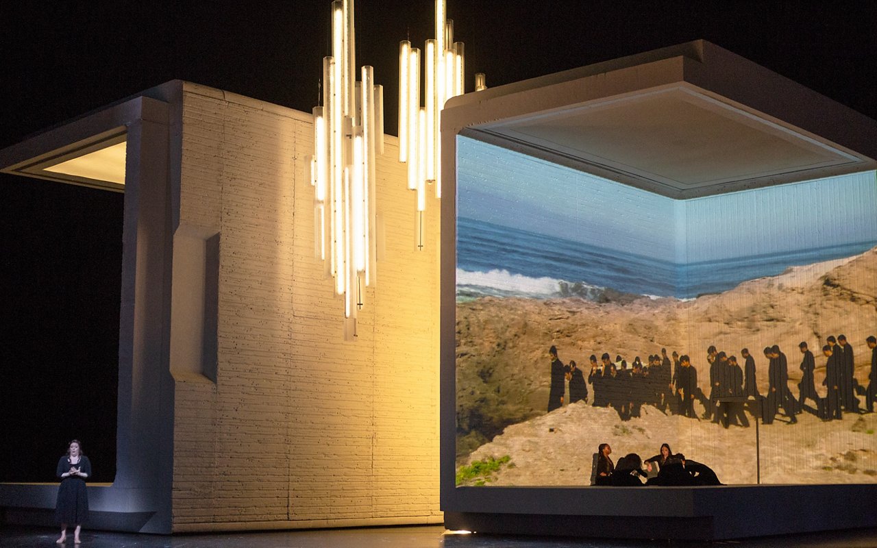 Theatrical scene with large architectural structures, a suspended installation of vertical lights and a projection of an arid landscape with a group of human figures in the background.