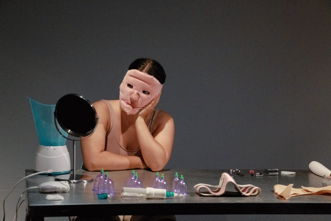 Persona con una máscara facial de silicona sentada frente a una mesa con un espejo, pequeños recipientes y diversos objetos, con la cabeza apoyada sobre una mano.