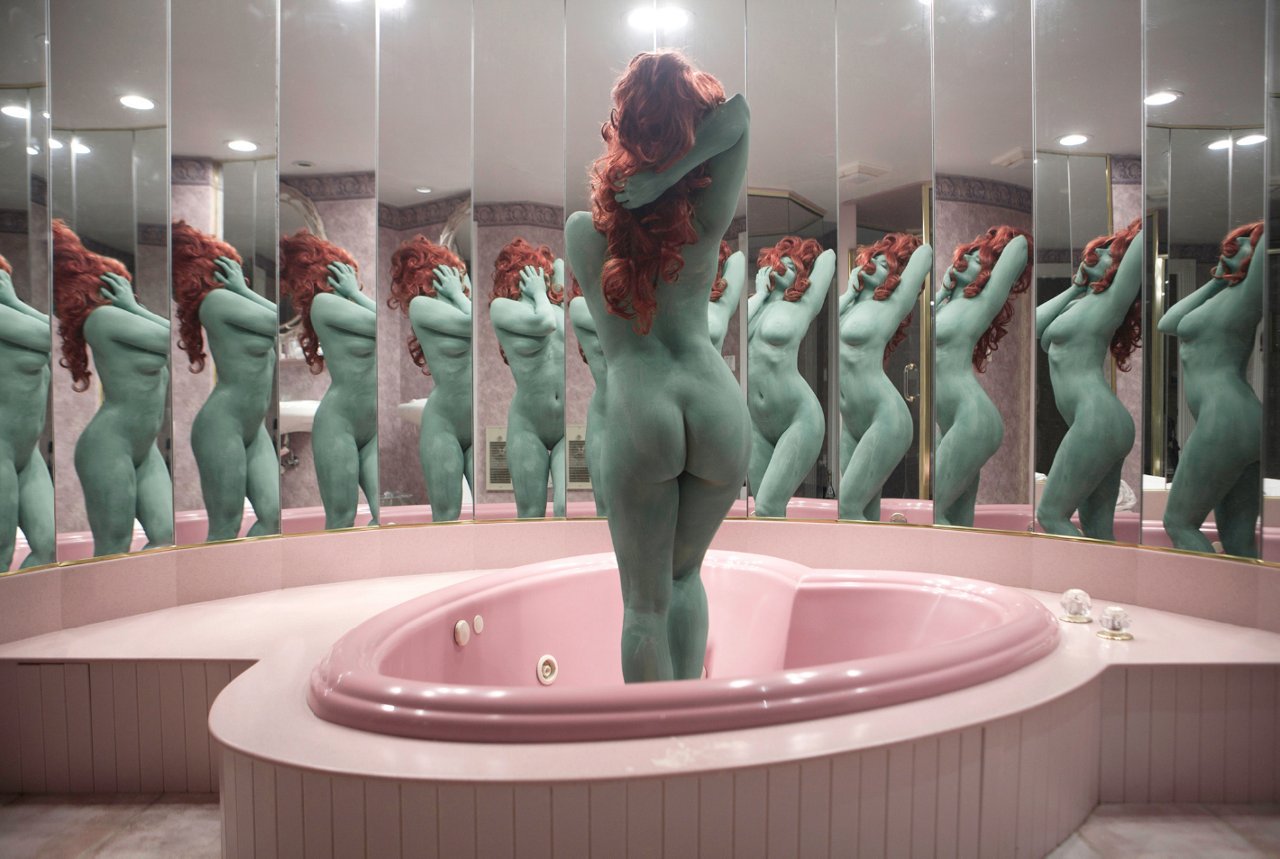 Chica desnuda con el cuerpo pintado de verde y el pelo rojo, posando de pie dentro de una bañera rosa con forma de corazón y rodeada de espejos que multiplican su imagen.