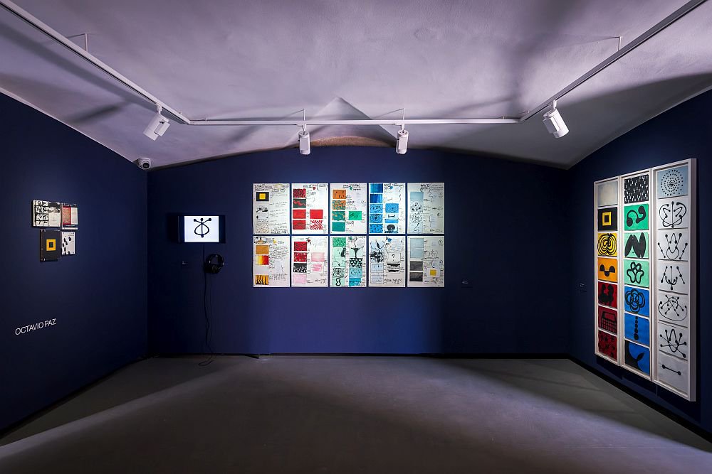 Sala de exposiciones de paredes oscuras con carteles y obras gráficas de colores dispuestos en cuadrículas, iluminados por focos, y una pequeña pantalla en una de las paredes.