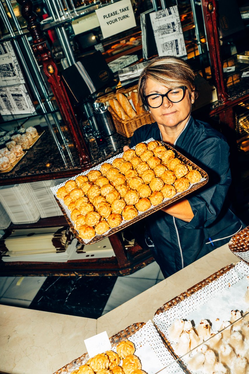 A pastry chef wearing glasses holds a tray of pine nut panellets behind the counter.