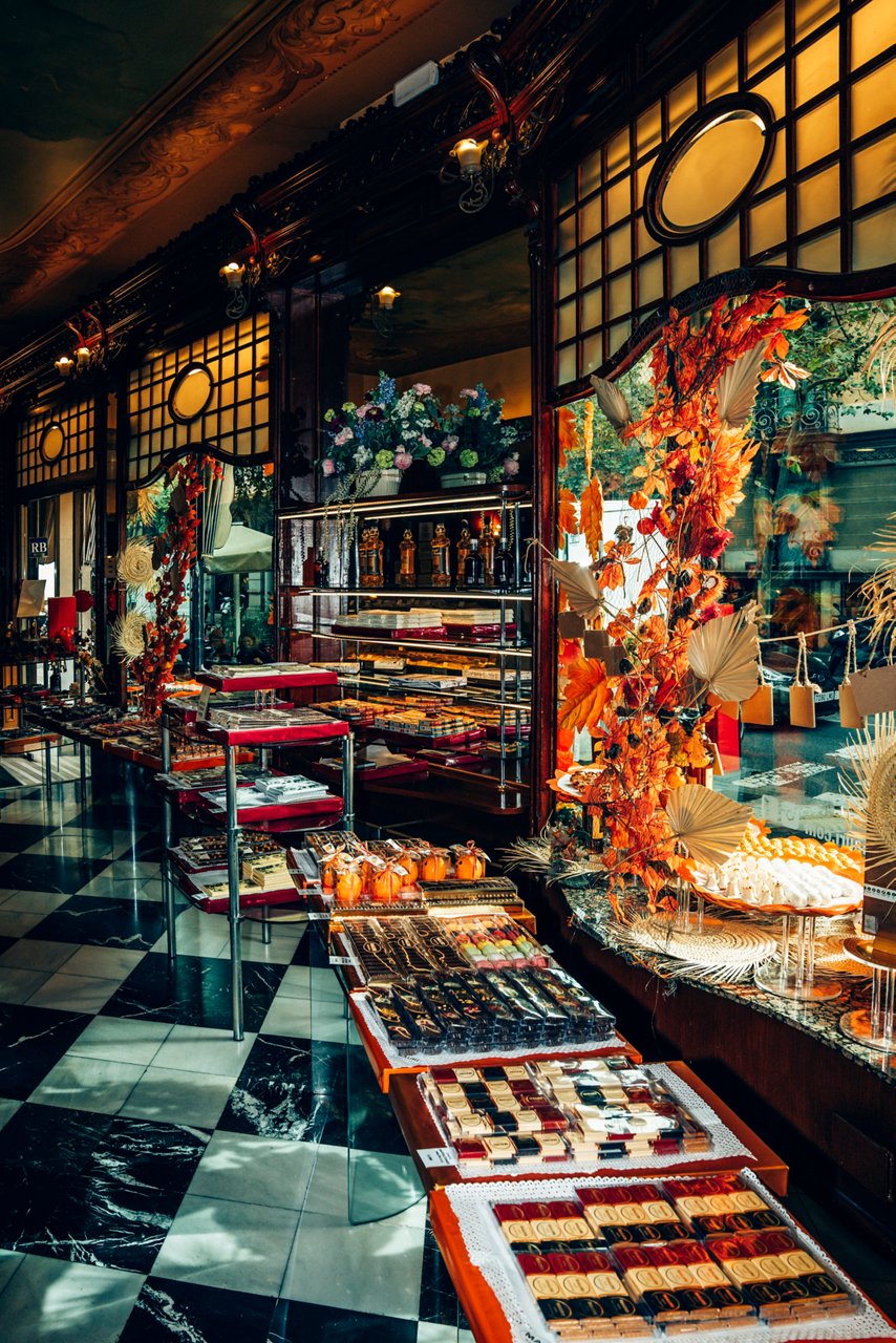 Inside a pastry shop with glass displays of sweets, floral decorations and a black and white chequered floor.