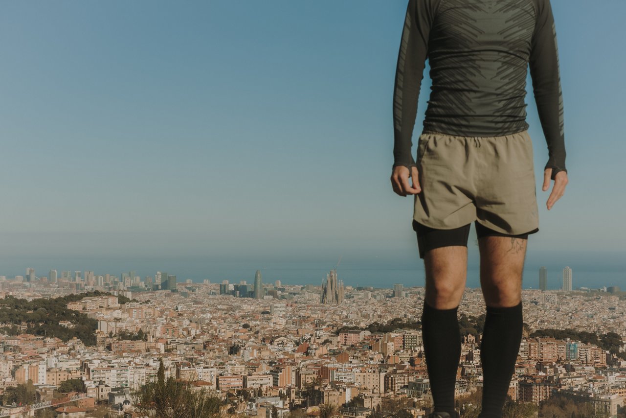 Home dempeus en una zona elevada de muntanya, amb una vista panoràmica de Barcelona, la Sagrada Família i el mar al fons.