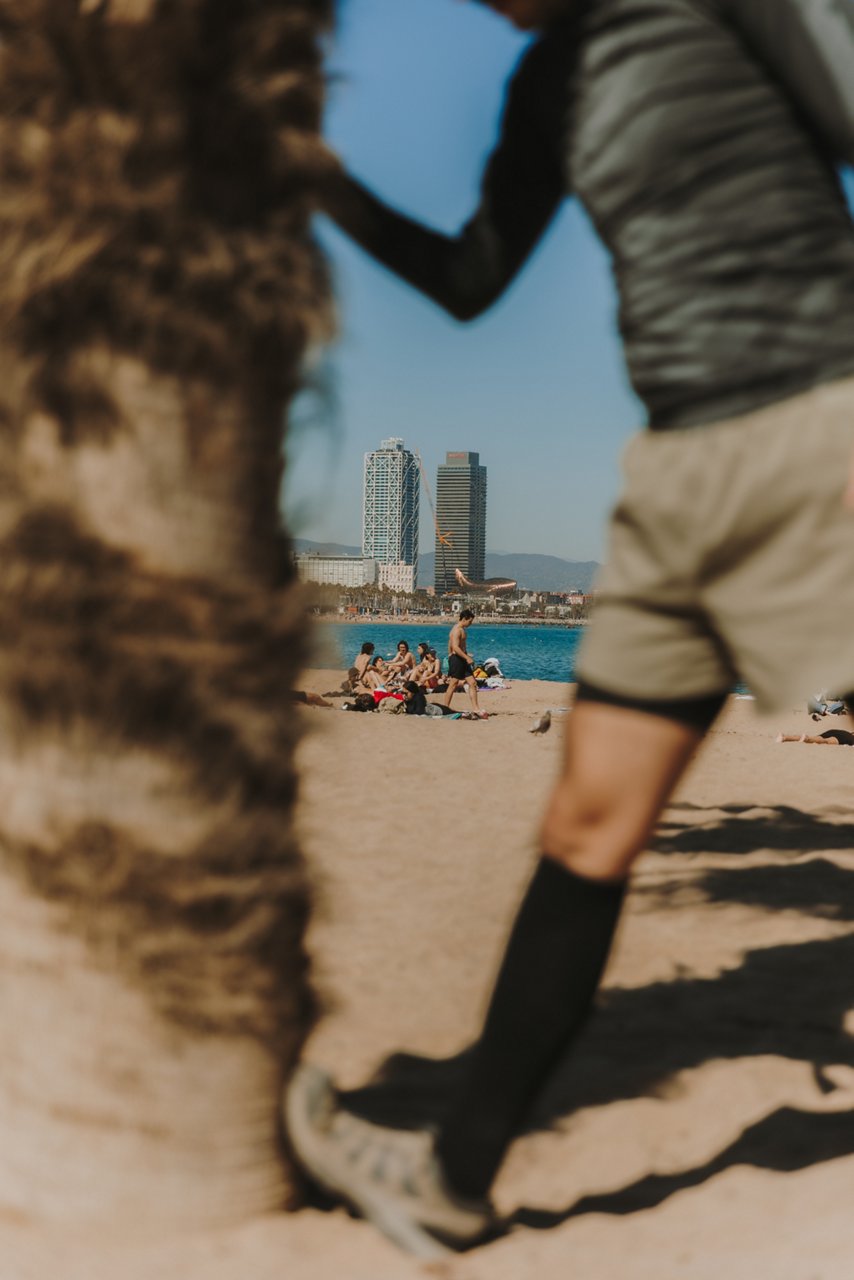 Persona fent estiraments a la platja al costat d’una palmera, amb mitjons de compressió. Al fons, banyistes, el mar i les torres del Port Olímpic. 