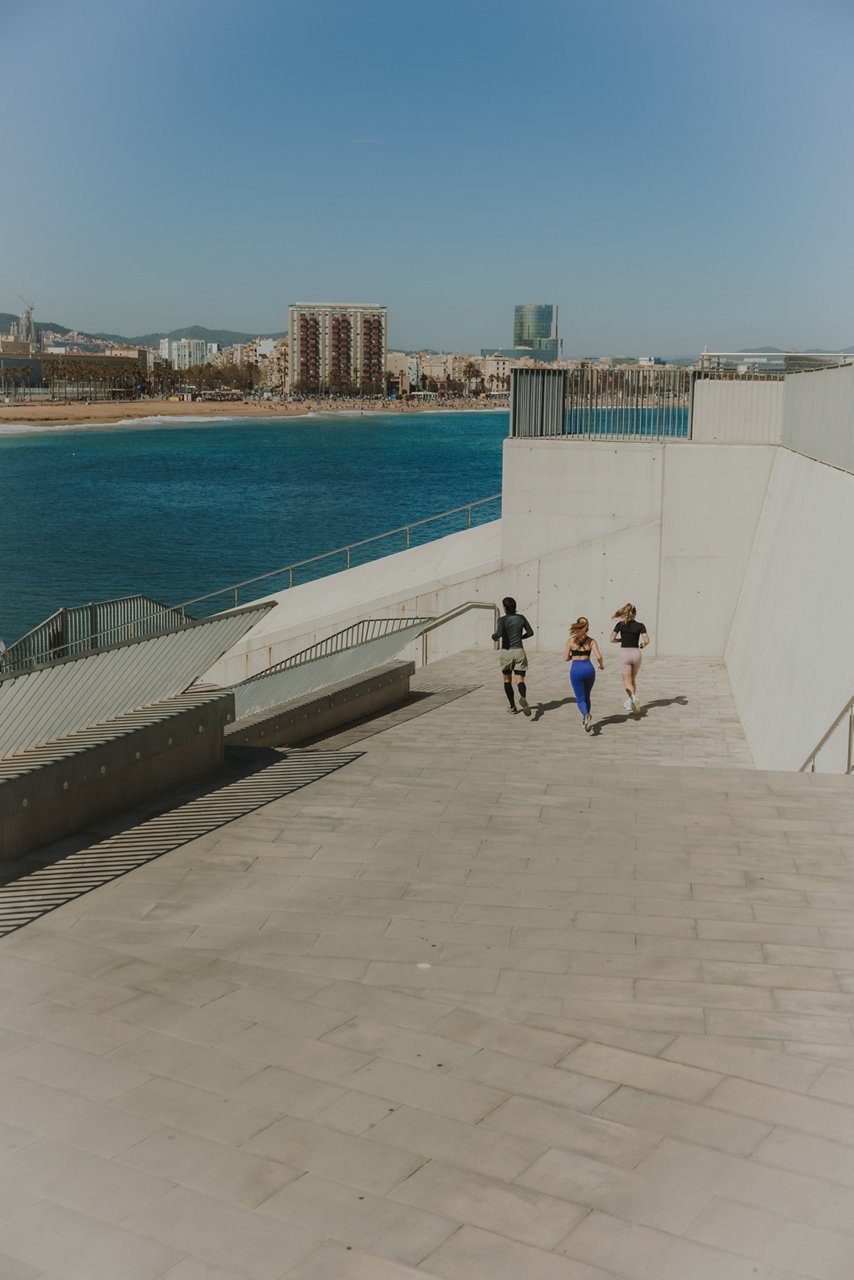 Tres persones corren per unes escales al costat del mar, amb la platja i els edificis de Barcelona al fons.