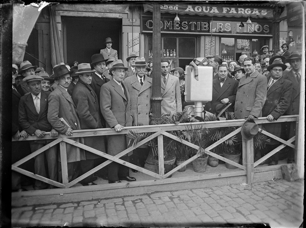 Photographie historique d’un grand groupe d’hommes réunis devant un établissement commercial. Tous portent de longs manteaux, des costumes formels et des chapeaux d’époque, regardant la caméra avec des expressions sérieuses ou curieuses. Ils se trouvent derrière une clôture en bois avec des pots de plantes à sa base. Au centre se distingue un appareil technique monté sur un support, l’un des premiers feux de circulation électriques de Barcelone, dont l’inauguration constitue le centre de l’événement. À l’arrière-plan, on voit la façade du magasin avec les enseignes « COMESTIBLES FINOS » et « AGUA FARGAS », ainsi que des vitrines présentant des produits.