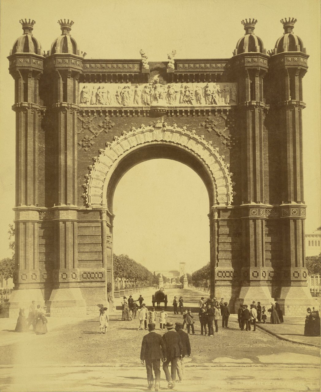 Fotografía histórica en color sepia del Arc de Triomf de Barcelona, una gran estructura monumental de ladrillo con un arco central ricamente decorado. El arco está flanqueado por torres con cúpulas ornamentadas y presenta relieves escultóricos en la parte superior con figuras humanas. Bajo el arco, varias personas vestidas con ropa de época pasean o se detienen, mientras al fondo se extiende un paseo arbolado que crea una profunda perspectiva. También se observa un carruaje en el centro. La escena corresponde a finales del siglo XIX, en el contexto de la Exposición Universal de Barcelona 1888, y transmite una atmósfera animada y urbana.