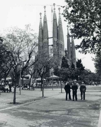Photographie historique en tons sépia de l’Arc de Triomf de Barcelone, une grande structure monumentale en brique avec un arc central richement décoré. L’arc est flanqué de tours aux coupoles ornées et présente des reliefs sculptés en partie supérieure représentant des figures humaines. Sous l’arc, plusieurs personnes vêtues d’habits d’époque se promènent ou s’arrêtent, tandis qu’à l’arrière-plan s’étend une promenade bordée d’arbres créant une perspective profonde. Une calèche est également visible au centre. La scène correspond à la fin du XIXe siècle, dans le contexte de l’Exposition universelle de Barcelone 1888, et transmet une atmosphère urbaine animée.