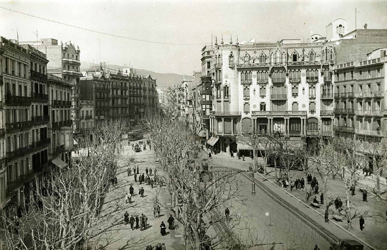 En blanc i negre, una avinguda ampla de Barcelona amb passeig central arbrat i edificis residencials als laterals. Els arbres, sense fulles, suggereixen una escena d’hivern. A la dreta destaca la Casa Fuster amb la façana ornamentada, balcons decoratius i grans obertures, que contrasta amb els edificis més sobris del voltant. Al centre del passeig hi ha vianants passejant, mentre que per la calçada circulen tramvies i alguns carruatges. L’escena mostra una ciutat activa, amb vida quotidiana als carrers i una composició que s’obre cap al fons, on es dibuixa el relleu de les muntanyes.