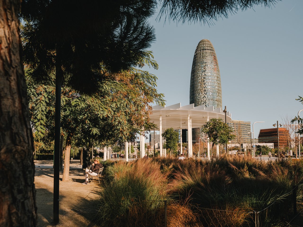View of Parc de les Glòries, Parc de les Glòries, with green areas and pathways, where a person sits on a bench under the trees. In the middle ground there is a modern pergola with white columns and a light roof. In the background rises the Torre Glòries, Torre Glòries, a tall cylindrical building with a glass façade that stands out over the urban surroundings. The scene depicts a calm, contemporary space, with vegetation and modern architecture coexisting in a sunny atmosphere.