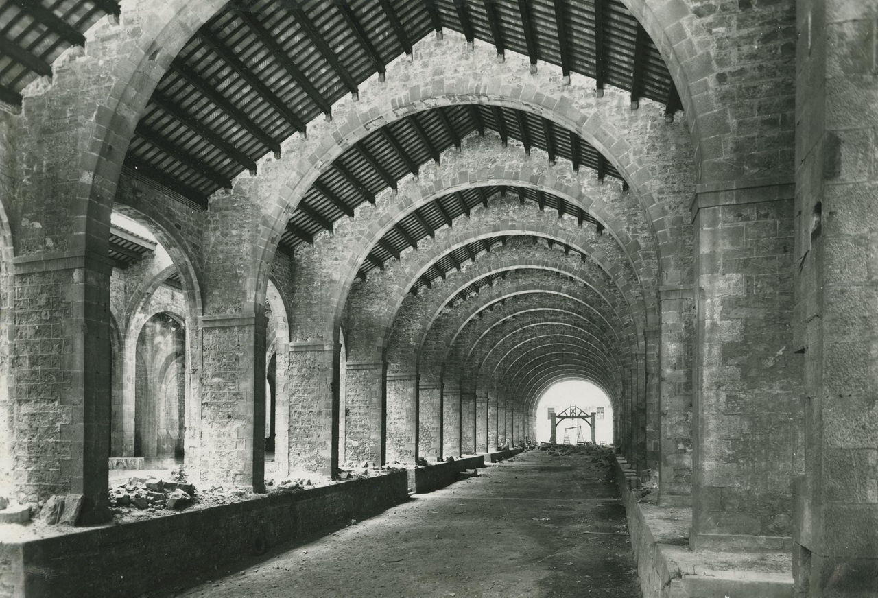 Foto en blanco y negro de las Drassanes Reials de Barcelona. Una gran nave cubierta con una sucesión de arcos de piedra de medio punto que se repiten rítmicamente hasta el fondo, creando una fuerte sensación de profundidad y simetría. La cubierta es de madera con vigas visibles, apoyada sobre los arcos. A ambos lados hay muros macizos de piedra con aberturas laterales. El suelo es de tierra batida o pavimento sencillo y el espacio aparece prácticamente vacío, con algunos elementos dispersos. Al fondo se ve una abertura que deja entrar la luz exterior, creando un núcleo de luz y cerrando la perspectiva.