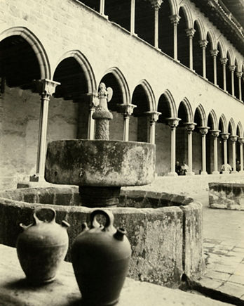 Fotografia en blanc i negre del claustre gòtic del monestir de Pedralbes, amb una galeria de dos nivells formada per arcs ogivals sostinguts per columnes esveltes. Els murs són de pedra i el conjunt presenta una gran regularitat i elegància. Al centre del pati hi ha una font circular de pedra amb una escultura d'un àngel al damunt. En primer terme es veuen gerres de ceràmica col·locades sobre un mur baix. A la galeria inferior, algunes persones passegen o seuen.