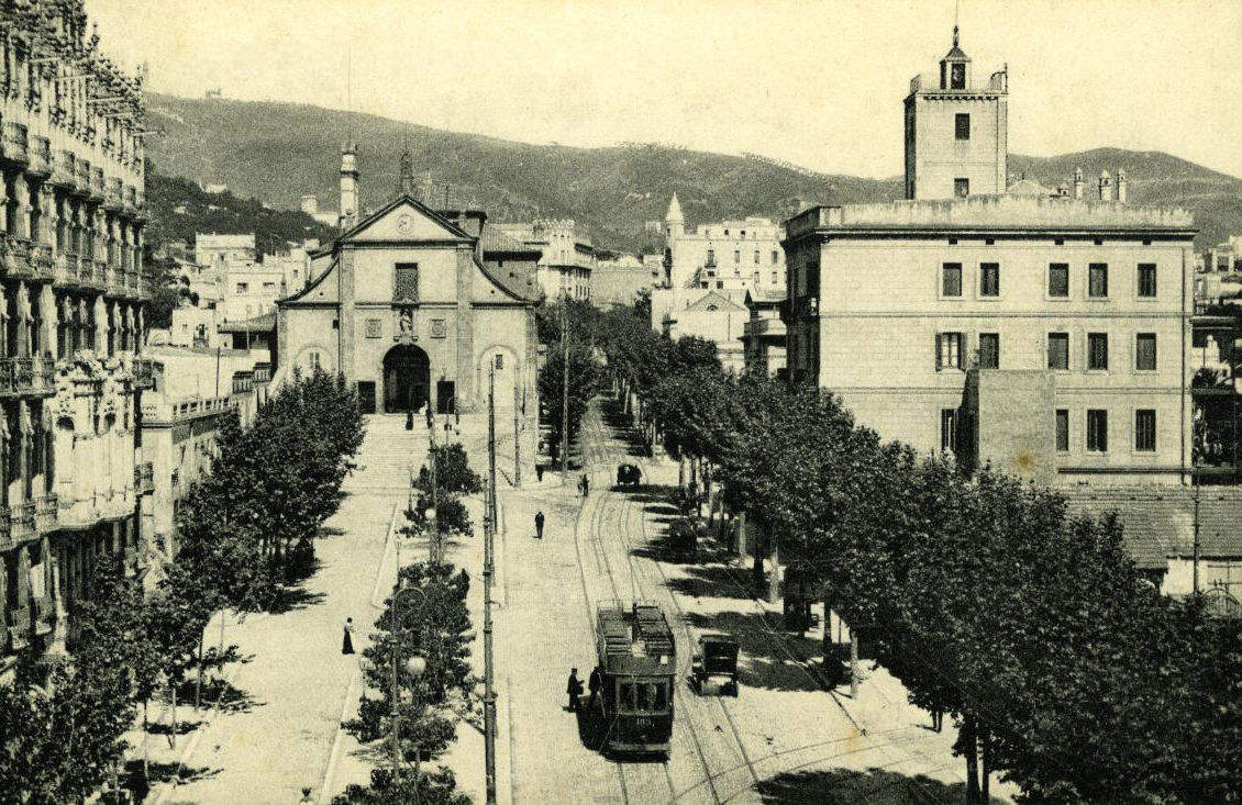 Vista urbana històrica d’un carrer ample i arbrat de Barcelona, amb una línia de tramvia circulant pel centre. A banda i banda hi ha fileres d’arbres que separen la via dels edificis, creant un passeig ordenat. Al fons destaca una església de façana senzilla amb portal central, mentre que a la dreta s’alça un edifici institucional amb torre. Es veuen alguns vianants i carruatges, i al centre un tramvia elèctric en circulació. Al fons, les muntanyes tanquen l’escena