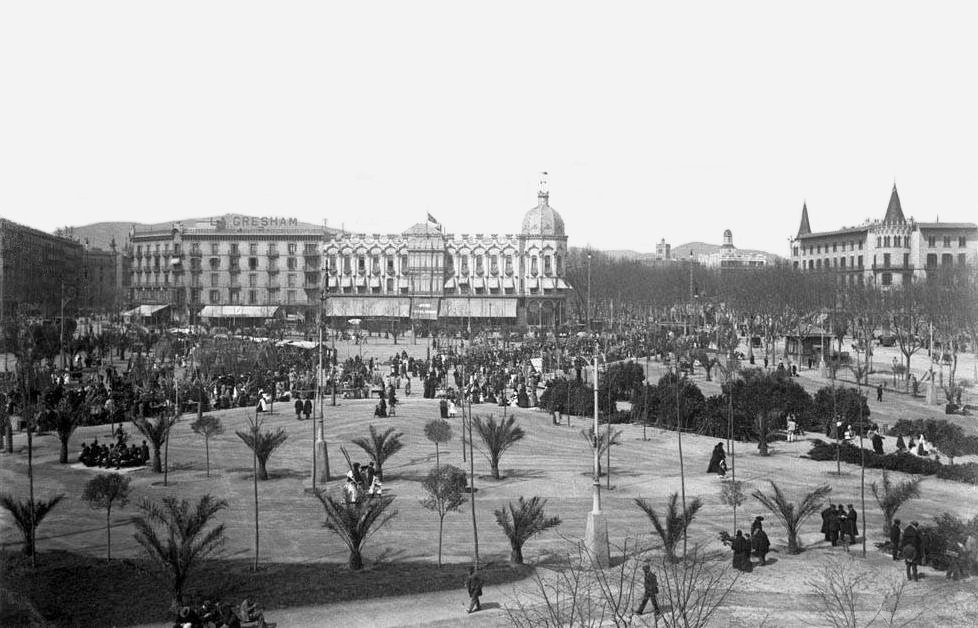 Fotografia històrica en blanc i negre de la plaça de Catalunya de Barcelona plena de gent passejant i reunida en grups, amb jardins i camins ordenats en primer terme. L’espai està urbanitzat amb arbres joves i zones verdes, i travessat per vials per a vianants. Al fons destaca un conjunt d’edificis emblemàtics: al centre, una construcció amb cúpula i façana ornamentada; a l’esquerra, un edifici amb el rètol “GRESHAM”; i a la dreta, edificis institucionals amb torres i teulades punxegudes. L’escena mostra una atmosfera animada i social, pròpia d’un espai públic central a inicis del segle XX.