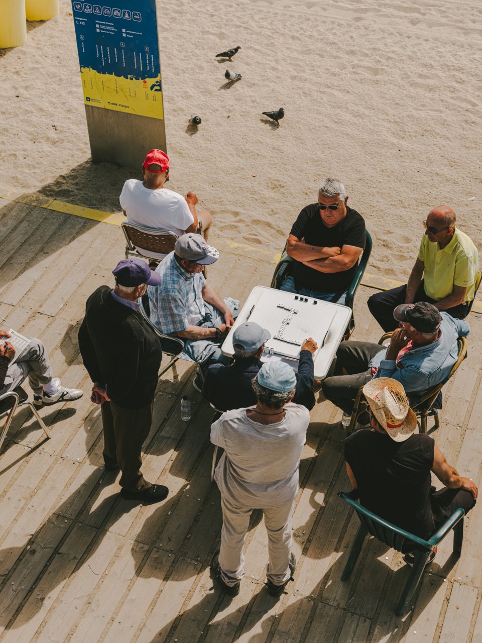 Group of people playing dominoes next to Barcelona beach, in a promenade area with sand and a few pigeons in the background.