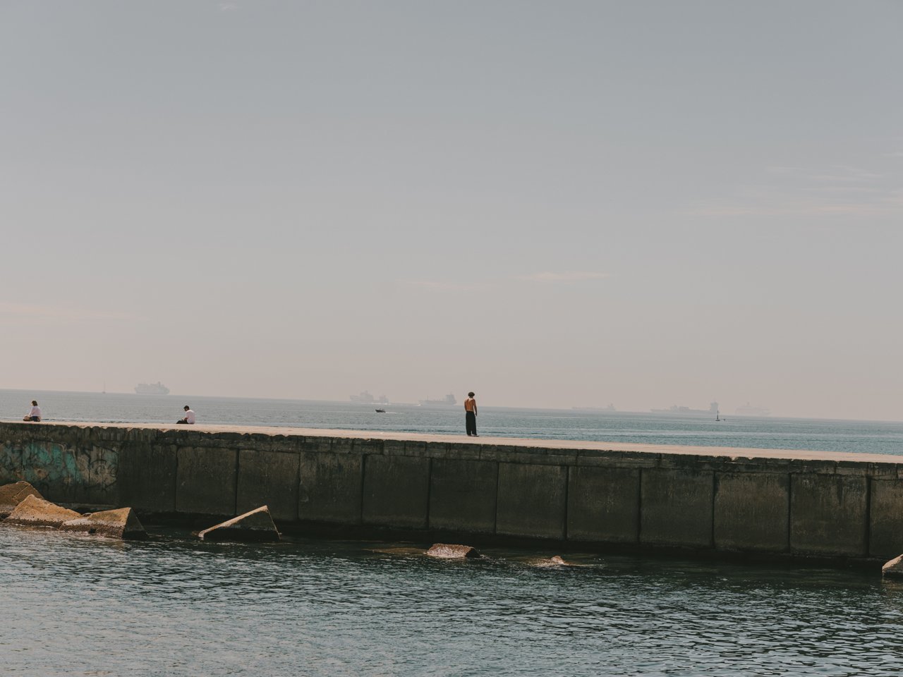 People walking and sitting on a breakwater by the sea in Barcelona, with ships on the horizon and calm waters in the foreground.