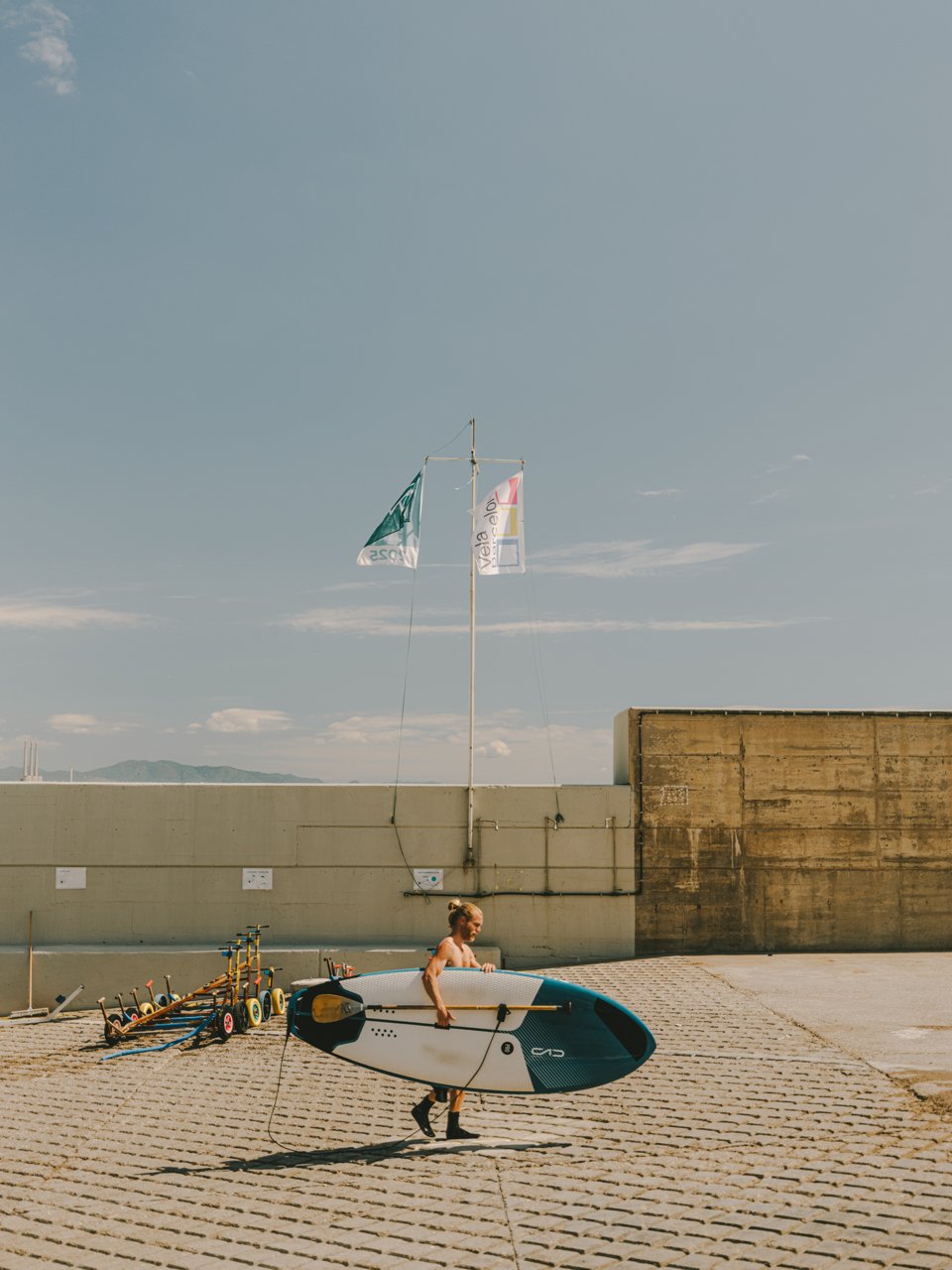 Person carrying a paddleboard in a paved area near the sea, with flags, paddles and the maritime horizon in the background.
