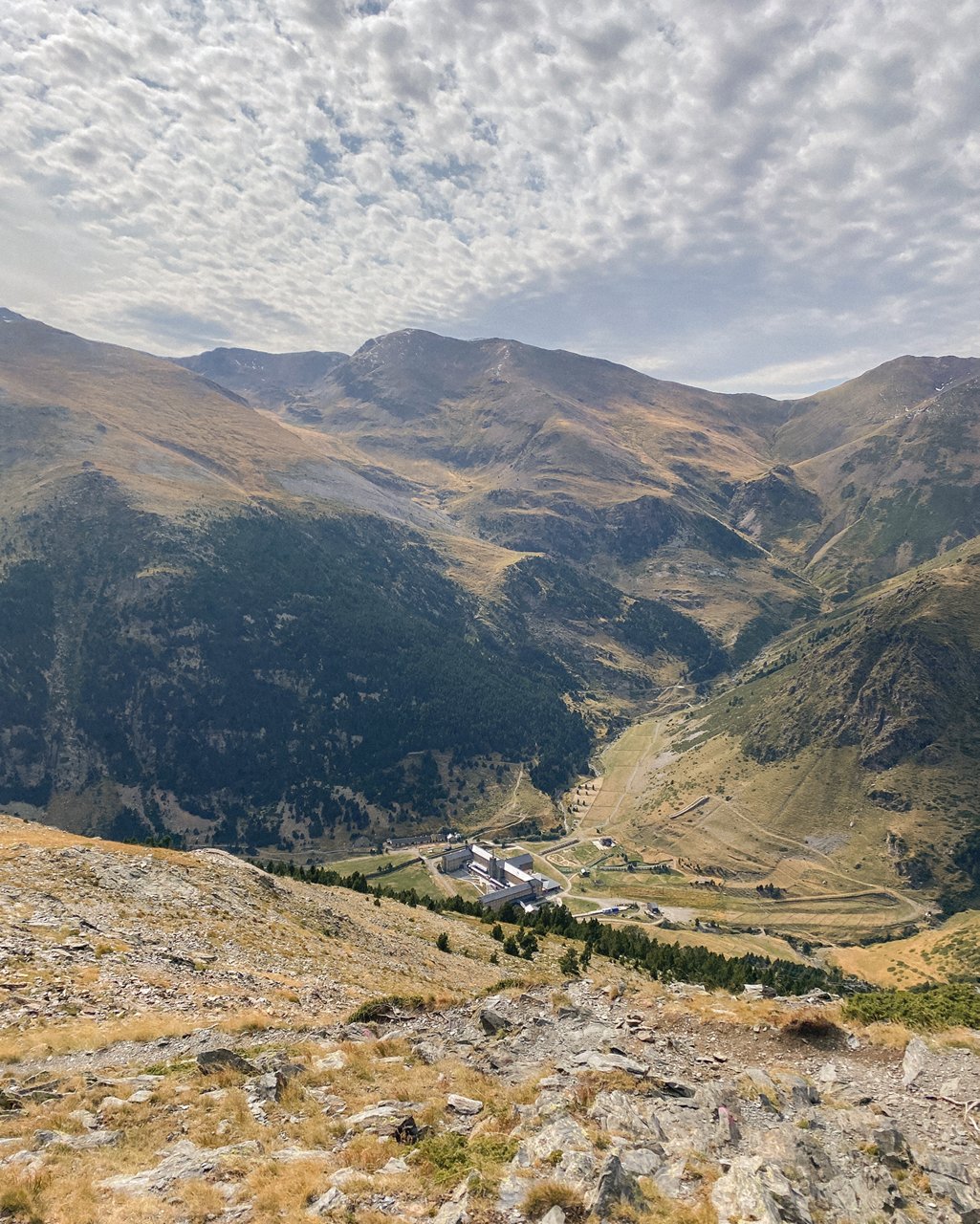 Vista panoramica di una valle montana con sentieri, edifici e rilievo ondulato sotto un cielo nuvoloso.