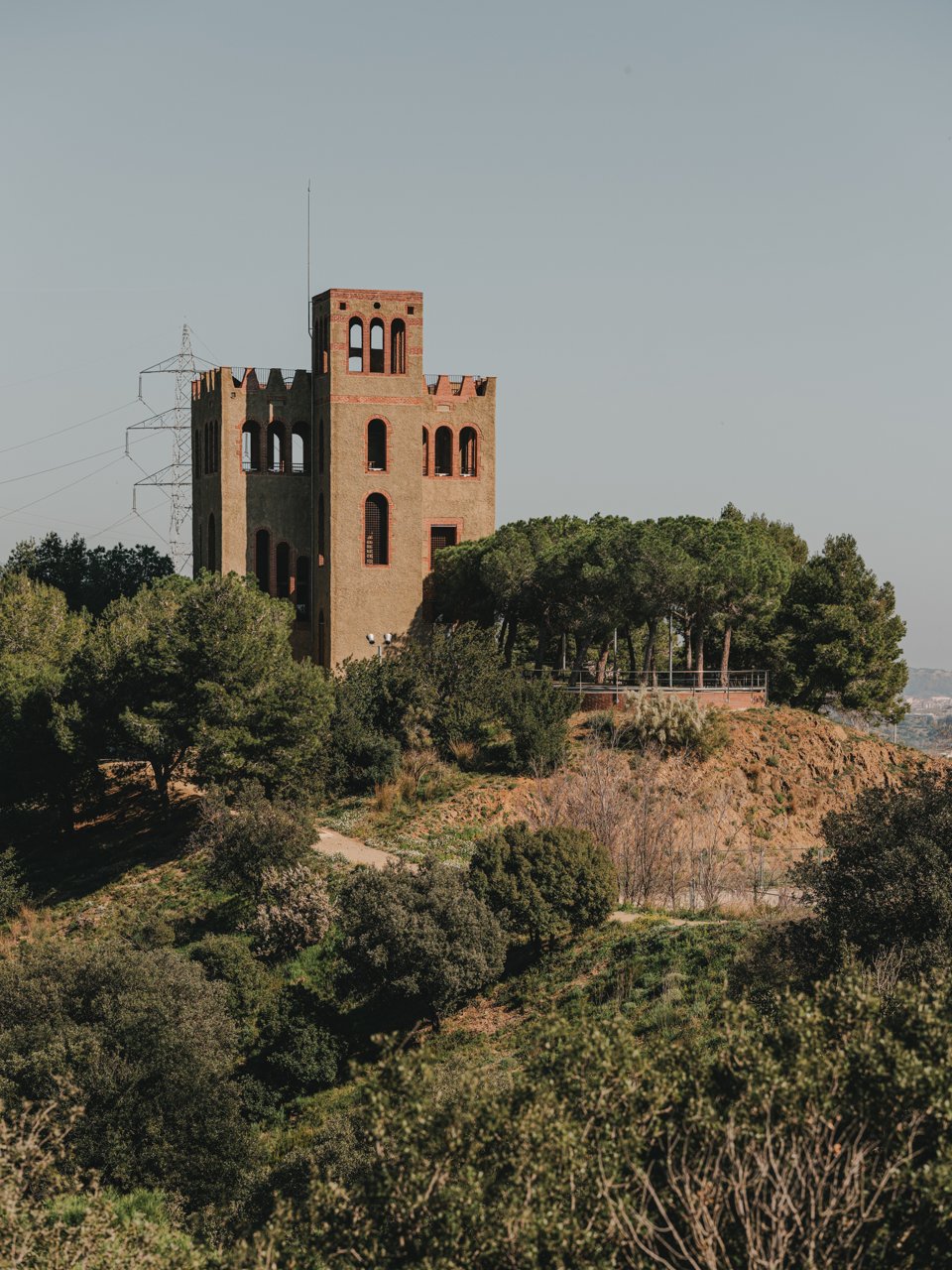 Torre de guaita de maó situada en un turó de Collserola, envoltada de pins i vegetació mediterrània, amb una línia elèctrica al fons. 