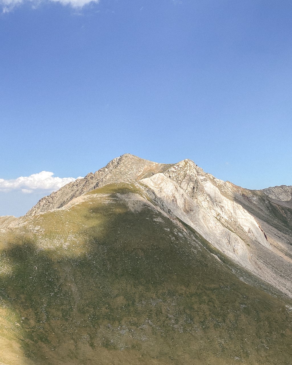 Cima de montaña con laderas herbosas y rocosas bajo un cielo azul con nubes dispersas.