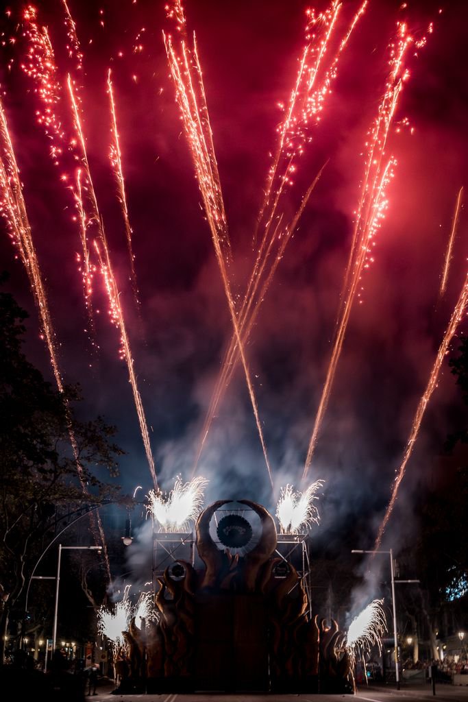Night-time pyrotechnic show featuring a large stage structure surrounded by fire, smoke and red sparks.