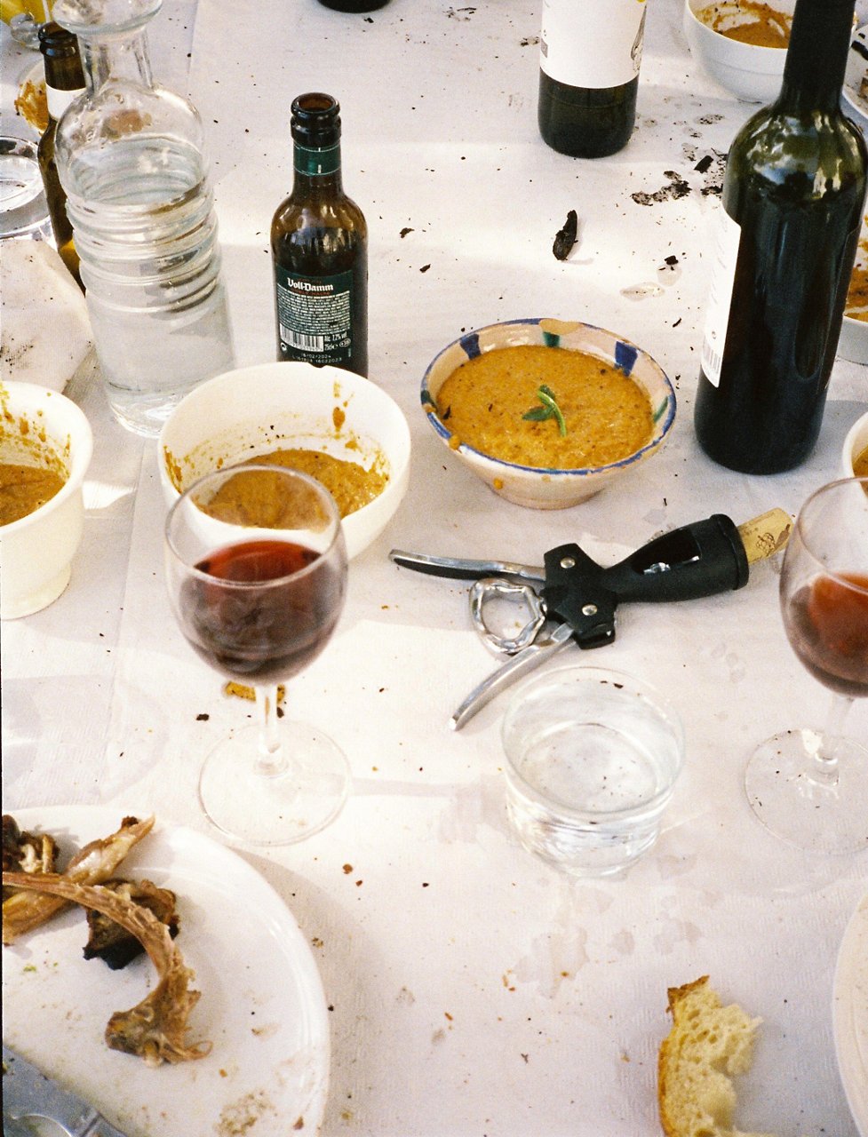 Détail d’une table après un repas, avec des verres de vin à moitié pleins, des bouteilles, des assiettes avec des restes de nourriture et un tire-bouchon au centre. Il y a aussi un bol de sauce, des verres d’eau et des miettes de pain éparpillées sur les nappes blanches, illustrant une ambiance informelle après le repas.