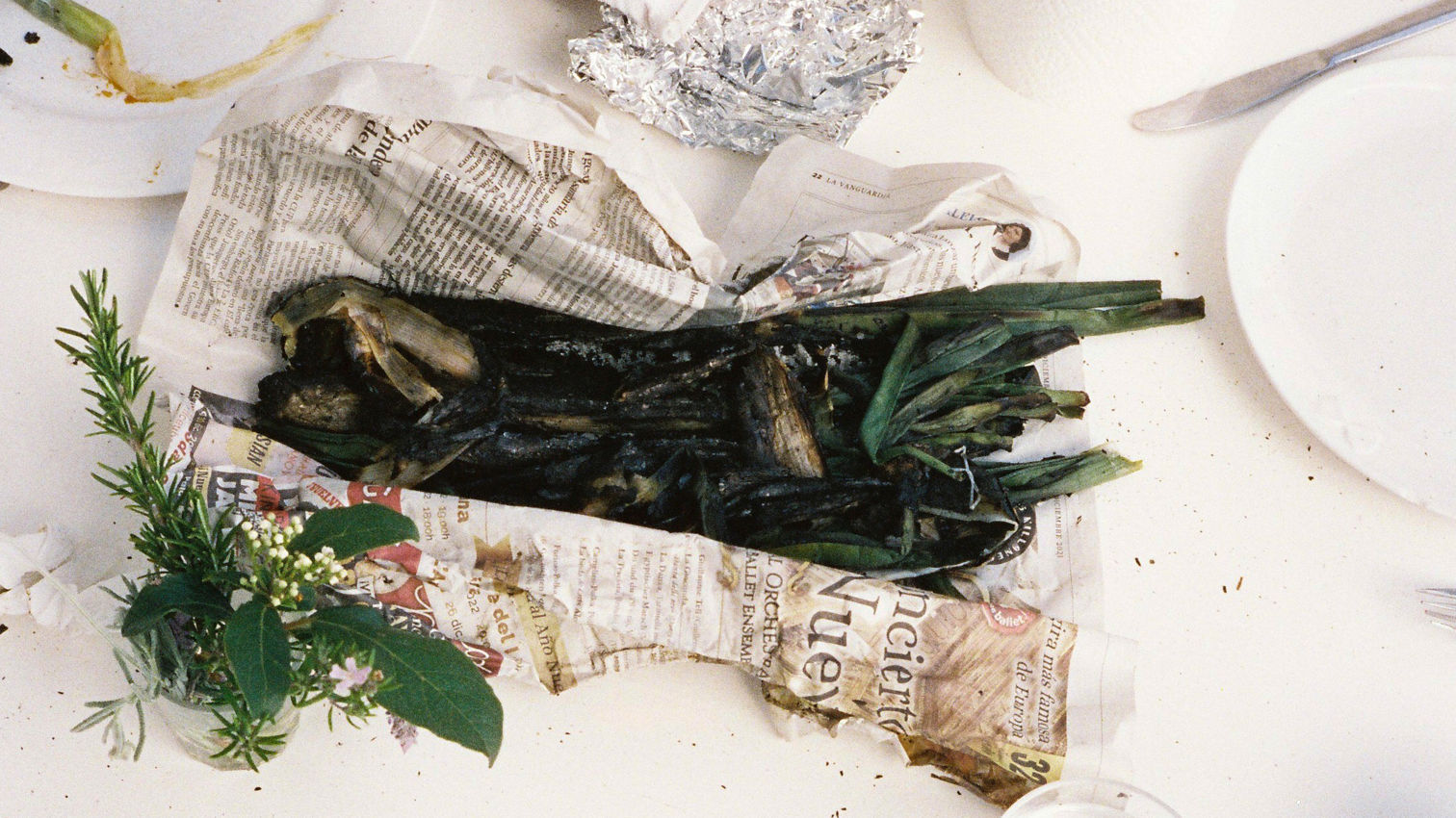 Manat de calçots sobre paper de diari, sobre una taula parada amb estovalles blanques. Al voltant, plats bruts i un rotlle de paper de cuina.