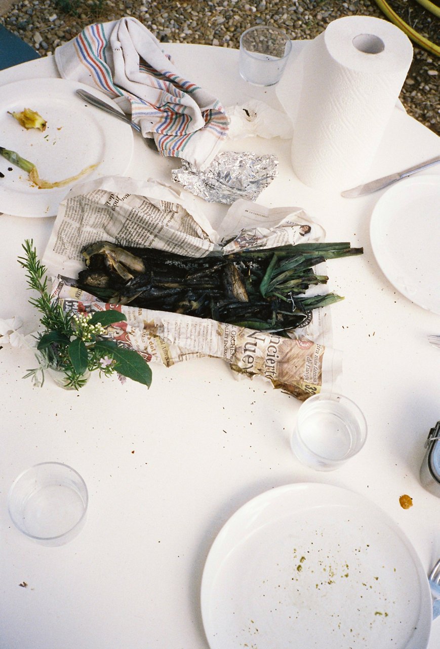 Mazzo di calçots su carta di giornale, su una tavola apparecchiata con tovaglie bianche. Intorno, piatti sporchi e un rotolo di carta da cucina.
