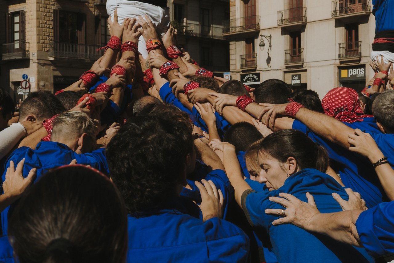 Un gruppo di persone con camicie blu alza le braccia e si tiene per mano, formando una struttura collettiva al centro di una piazza. Alcuni indossano fazzoletti rossi al polso, e la scena trasmette coordinazione, forza e spirito di squadra, in un contesto urbano con edifici sullo sfondo.