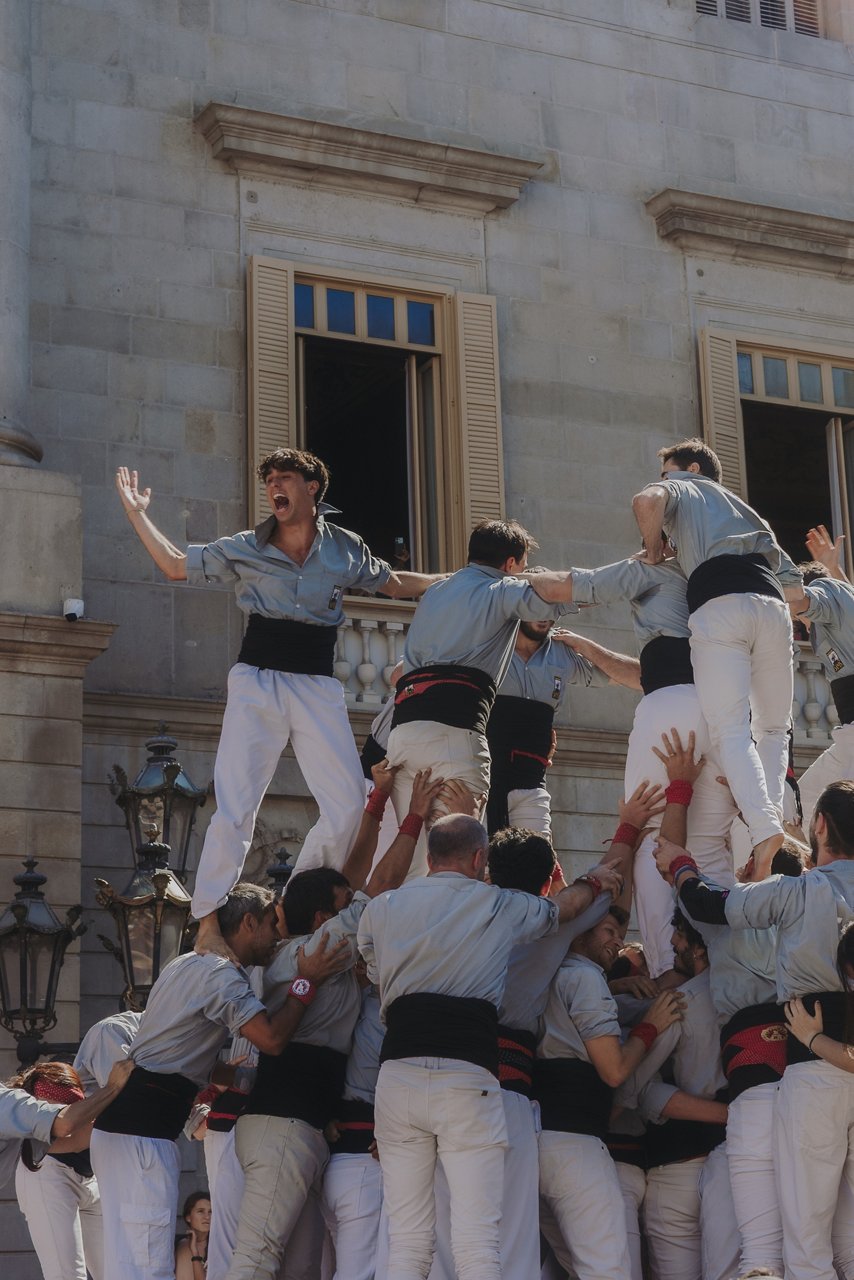 Grup de joves vestits amb camisa gris clar i faixa negra estan acabant de descarregar un castell davant d’un edifici a la plaça Sant Jaume. Un d’ells crida amb el braç aixecat, celebrant l’èxit d’haver pogut carregar i descarregar el castell, mentre els altres s’agafen per mantenir l’equilibri.